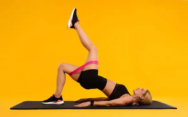 Woman exercising with elastic resistance band on fitness mat against orange background