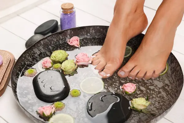 Woman soaking her feet in plate with water, stones, flowers and lime slices on white wooden floor, closeup. Pedicure procedure