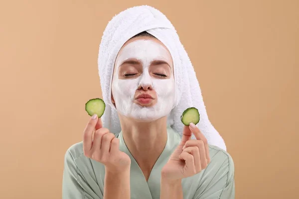 Woman with face mask and cucumber slices sending air kiss on beige background. Spa treatments