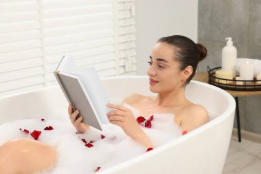 Küvette köpük ve gül yapraklarıyla banyo yapan kadın kitap okuyor.