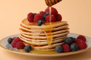 Soluk turuncu arka planda ahududu ve yaban mersinli leziz krep yığınının üzerine bal dökülüyor.