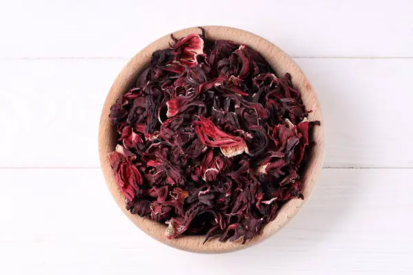 Dry hibiscus tea in bowl on white wooden table, top view