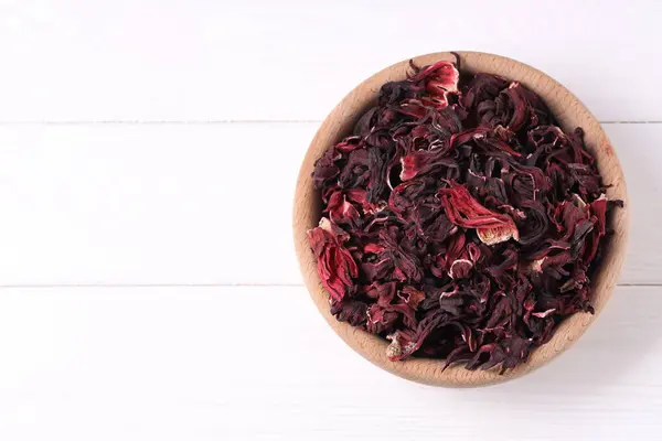 Dry hibiscus tea in bowl on white wooden table, top view. Space for text