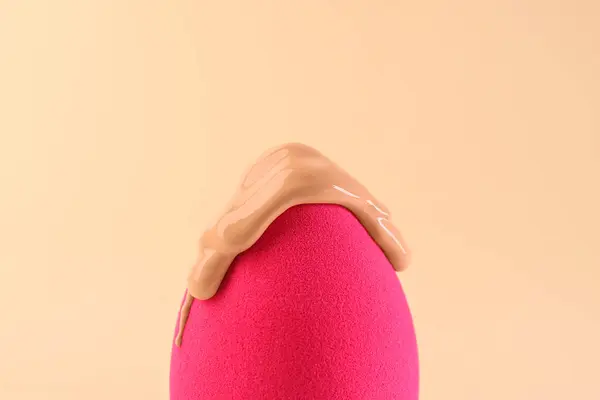 Pink makeup sponge with skin foundation on beige background, closeup