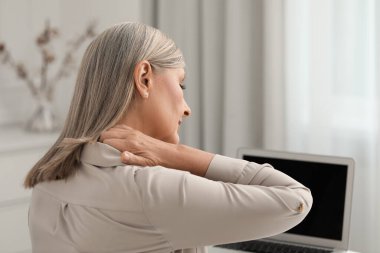 Woman suffering from neck pain at workplace in room clipart