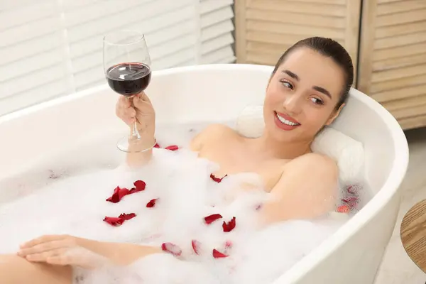 Happy woman with glass of wine taking bath in tub with foam and rose petals indoors