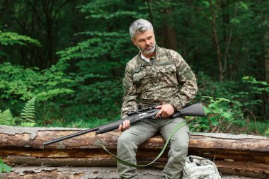 Avcı tüfekli bir adam ormanda devrilmiş ağaçta oturuyor. Metin için boşluk