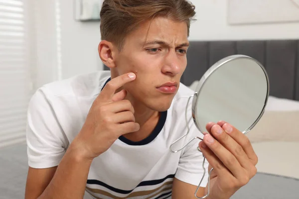 Upset young man looking at mirror and touching pimple on his face indoors. Acne problem