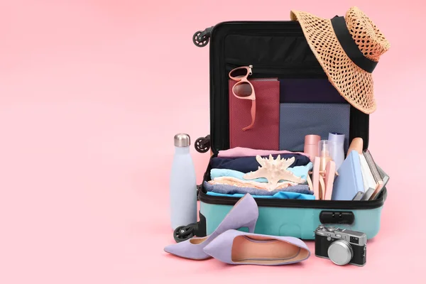 Open suitcase with clothes, beach accessories and shoes on pink background, space for text. Summer vacation