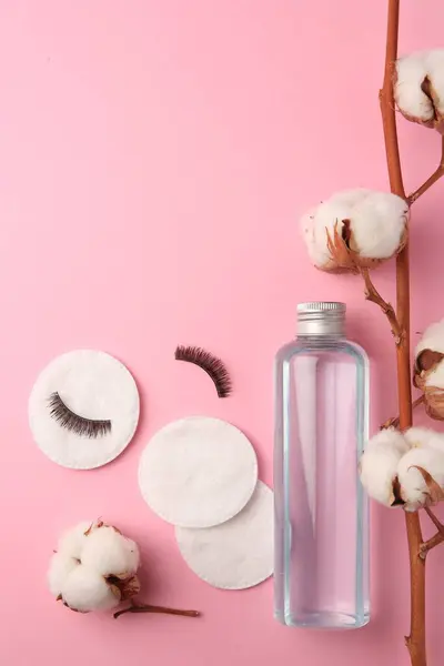 Bottle of makeup remover, cotton flowers, pads and false eyelashes on pink background, flat lay. Space for text