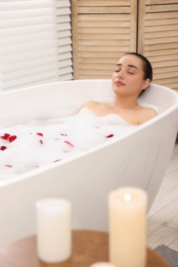 Kadın küvette köpük ve gül yapraklarıyla banyo yapıyor.