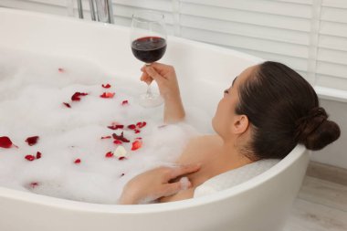 Elinde şarap bardağıyla küvette köpük ve gül yapraklarıyla banyo yapan bir kadın.