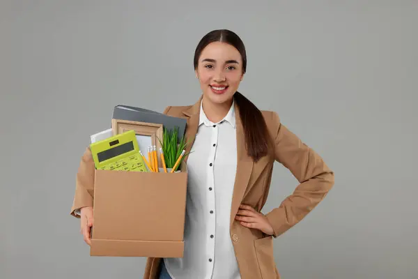 Happy unemployed woman holding box with personal office belongings on grey background