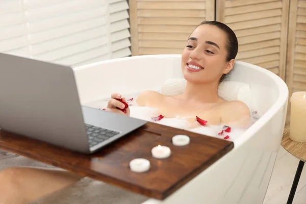 Happy woman with laptop taking bath in tub with foam and rose petals indoors