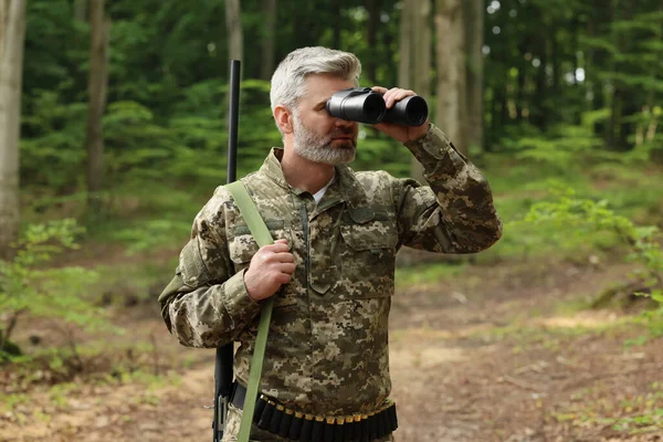 Man with hunting rifle looking through binoculars in forest