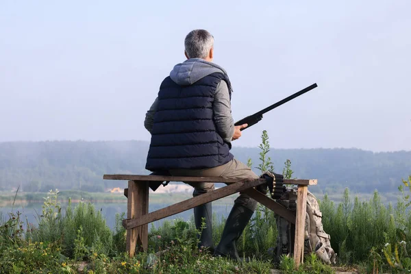 Man with hunting rifle sitting on wooden bench near lake outdoors, back view