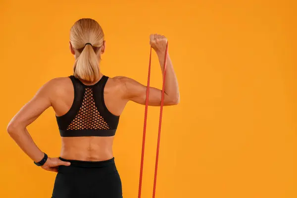 Woman exercising with elastic resistance band on orange background, back view. Space for text
