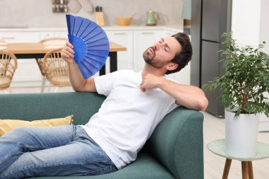 Sakallı adam evdeki kanepede serinlemek için mavi el yelpaze sallıyor.