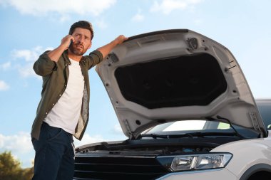 Telefondan konuşan üzgün bir adam yol kenarında bozuk bir arabanın yanında, düşük açılı bir manzara.