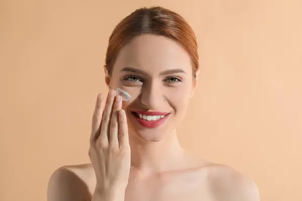Beautiful young woman with sun protection cream on her face against beige background