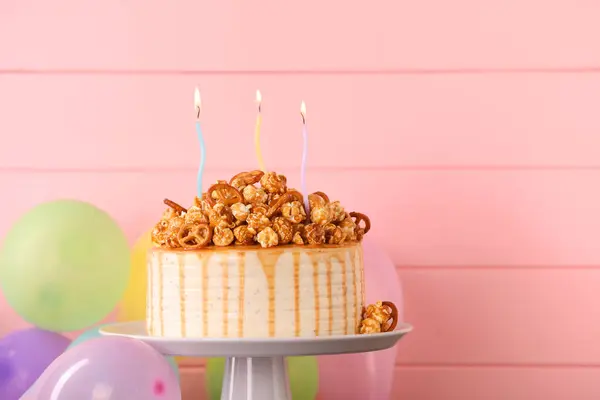 Caramel drip cake decorated with popcorn and pretzels near balloons against pink wooden background, space for text
