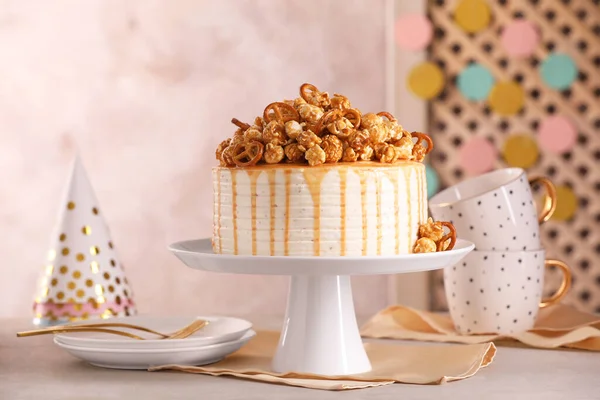 Caramel drip cake decorated with popcorn and pretzels near tableware on light grey table