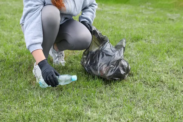 Woman with trash bag picking up plastic bottle outdoors, closeup. Recycling concept