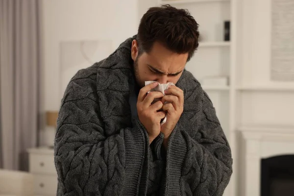 Sick man wrapped in blanket with tissue blowing nose at home. Cold symptoms