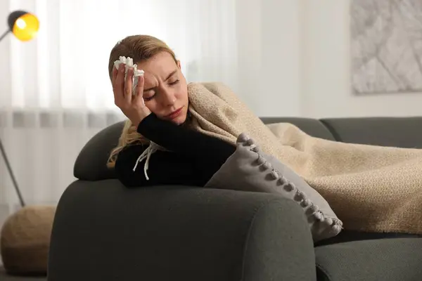Sick woman with tissue under blanket on sofa at home. Cold symptoms