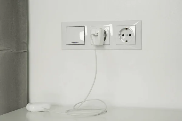 Charging case of modern wireless earphones plugged into power socket on white table indoors