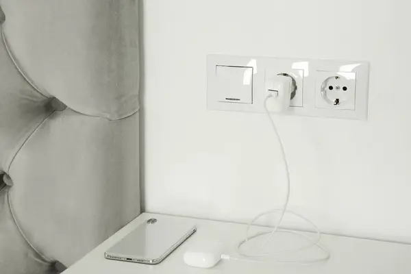 Charging case of modern wireless earphones plugged into power socket and smartphone on white table indoors
