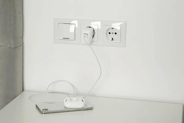Charging case of modern wireless earphones plugged into power socket and smartphone on white table indoors
