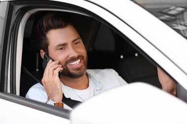 Mutlu sakallı adam arabada akıllı telefondan konuşuyor, dışarıdan bakıyor.