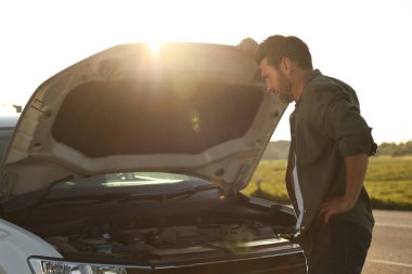 Kırık bir arabanın kaputunun altına bakan üzgün bir adam.