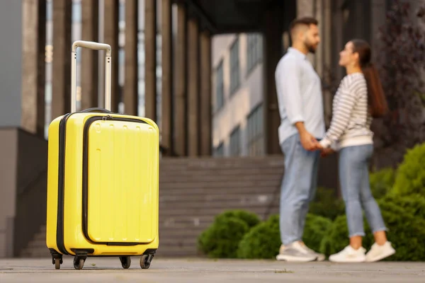 Long-distance relationship. Young couple holding hands near building outdoors, focus on yellow suitcase