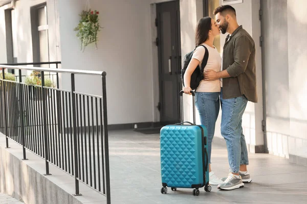 Long-distance relationship. Beautiful young couple with luggage kissing near house entrance outdoors