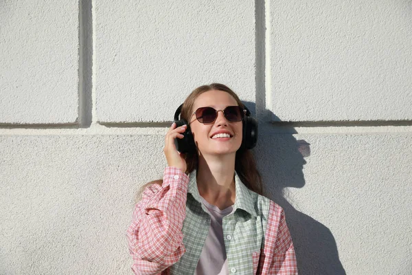 Smiling woman in headphones listening to music near white wall outdoors