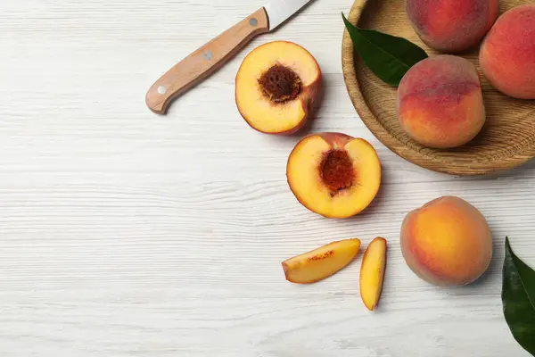 Delicious juicy peaches, leaves and knife on white wooden table, flat lay. Space for text