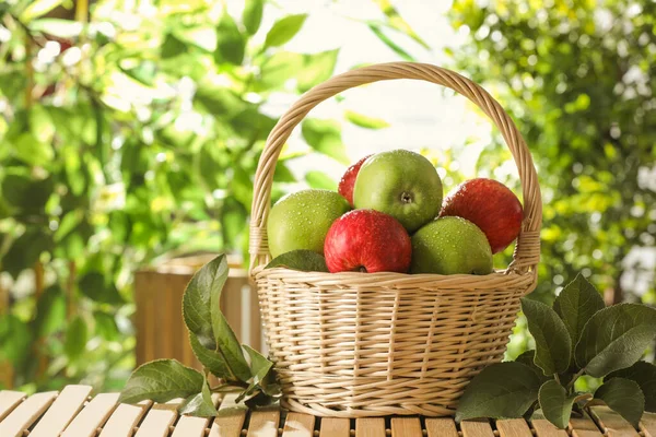 Different wet apples in wicker basket and green leaves on wooden table outdoors