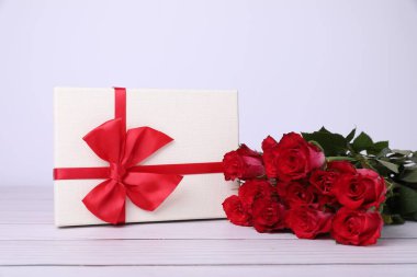 Kırmızı fiyonklu hediye kutusu ve beyaz ahşap masada güzel güller.
