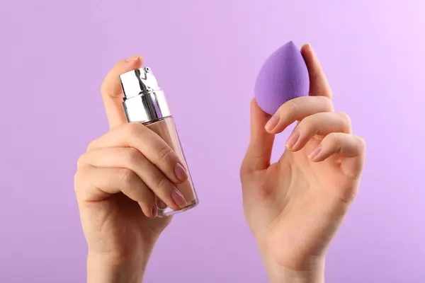 Woman with makeup sponge and skin foundation on violet background, closeup