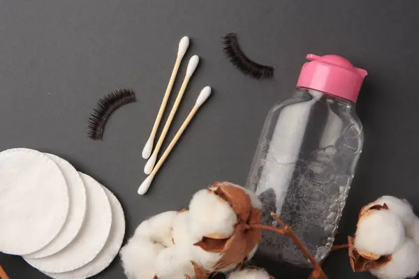 Bottle of makeup remover, cotton flowers, pads, swabs and false eyelashes on grey background, flat lay