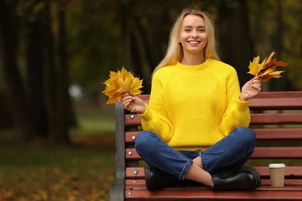 Happy woman with autumn leaves on bench in park. Space for text