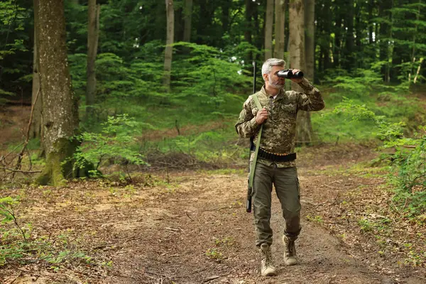 Man with hunting rifle looking through binoculars in forest. Space for text