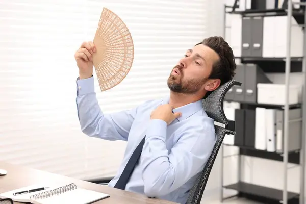 Bearded businessman waving hand fan to cool himself at table in office