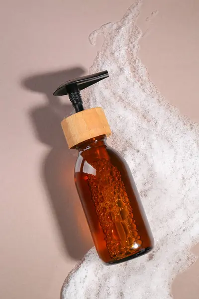 Bottle of cosmetic product and foam on brown background, top view