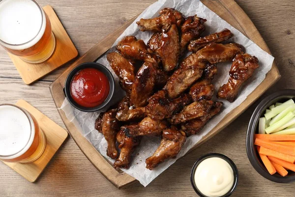 Tasty chicken wings, glasses of beer, sauces and vegetable sticks on wooden table, flat lay. Delicious snacks
