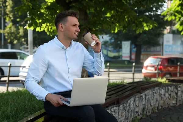 Handsome man with laptop drinking coffee on bench outdoors. Space for text
