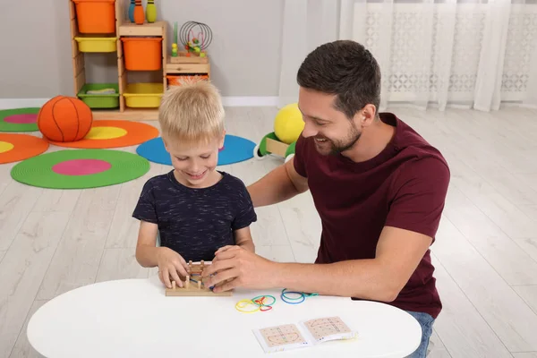 Motor skills development. Happy father helping his son to play with geoboard and rubber bands at white table in room