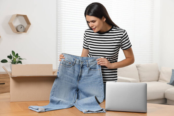 Молодая женщина с только что распакованными джинсами, картонной коробкой и ноутбуком на столе в помещении. Интернет-магазины